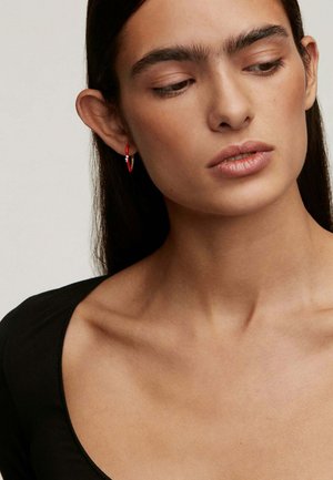 Aro rojo con acento plateado, diseño ligero y textura suave. Lleva puesto en la oreja izquierda sobre un fondo neutro.