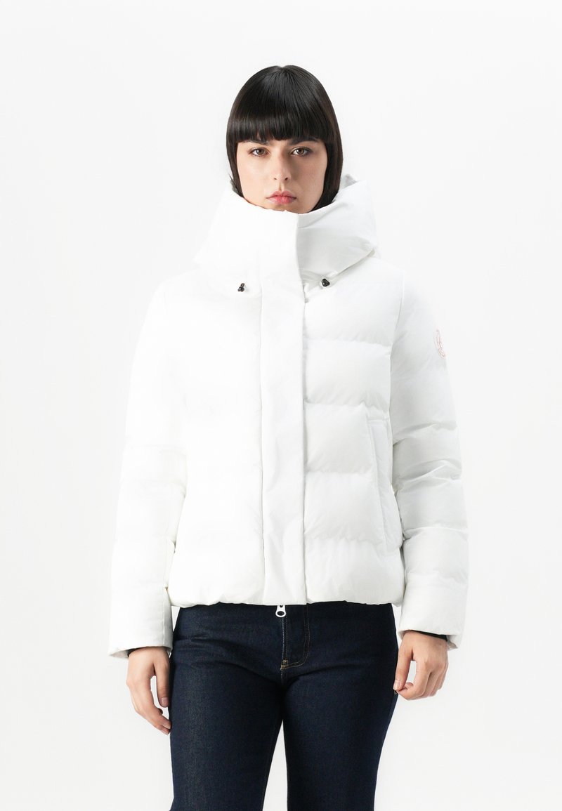 Giacca a vento bianca con colletto alto, chiusure a pressione, maniche corte e una texture trapuntata. Indossata sopra jeans scuri.