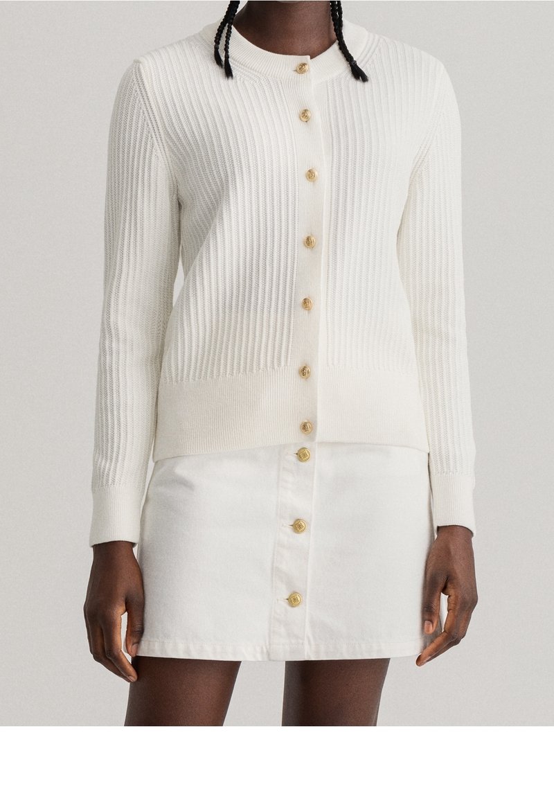 Bílý vroubkovaný cardigan se zlatými knoflíky, s kulatým výstřihem a upnutými rukávy. Spárován s bílou denimovou sukní se shodnými knoflíky.