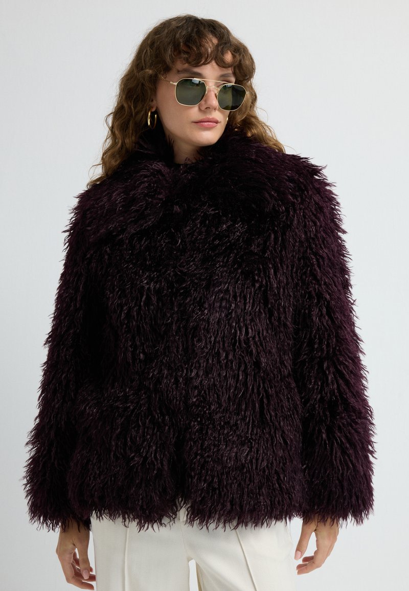 Kvinde iført en mørk lilla fluffy faux fur jakke, hvide bukser, gyldne hoops øreringe og grønne aviator solbriller mod en ensfarvet baggrund.