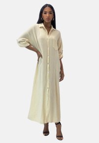 Lang, lysegul skjortekjole lavet af tekstureret stof. Har en enkelt brystlomme og opviklede ærmekanter. Båret med sorte sandaler.