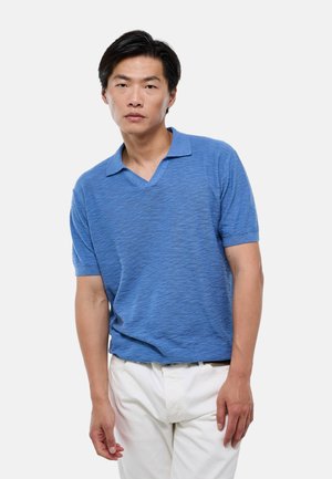 Homme adulte aux cheveux noirs portant une chemise bleue à manches courtes avec col et un pantalon blanc sur un fond blanc uni.