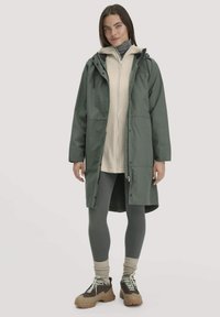 Frau in einer grünen Kapuzenparka über einem beigen Reißverschluss-Hoodie, dunklen grauen Leggings, beigen Socken und braunen Wanderschuhen, die vor einem neutralen Hintergrund steht.