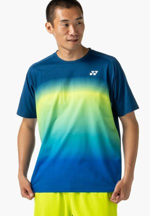 Uomo che indossa una maglietta sportiva a gradiente blu con logo bianco sul petto e pantaloncini giallo brillante, che tiene i lati della maglietta e guarda a destra.