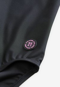 Maillot de bain noir en matériau nylon texturé, avec une finition lisse et un patch logo circulaire avec des lettres roses sur le côté gauche.