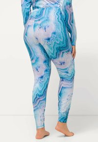 Primo piano di una persona che indossa leggings blu e bianchi con motivo a marmo e un top abbinato, in piedi a piedi nudi su uno sfondo bianco uniforme.