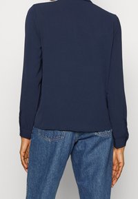 Mörkblå långärmad blus med krage och knäppte manschetter, tillverkad av slät tyg, tillsammans med högmidjade blå denimjeans.