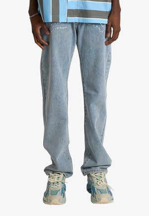 Persona che indossa jeans azzurri chiari con dettagli in strass sulle tasche anteriori e sneakers chunky blu-grigie, tagliati a vita e piedi.