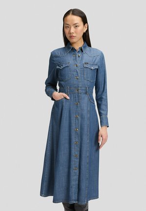 Frau trägt ein langärmeliges, blaues Jeanskleid mit Knopfleiste, Brusttaschen und Gürtelschlaufen, steht vor einem einfarbigen grauen Hintergrund.
