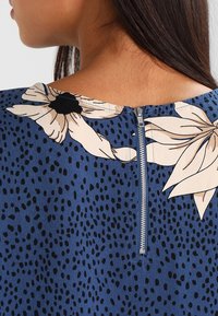 Blouse bleue à fleurs dotée d'une fermeture éclair au dos, avec un motif de grandes fleurs et de petits points noirs sur un fond uni.