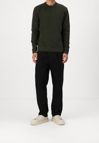 Tmavě zelený pletený svetr s kulatým výstřihem, doplněný o černé kalhoty s širokými nohavicemi a světle béžové tenisky s texturovanou podrážkou.