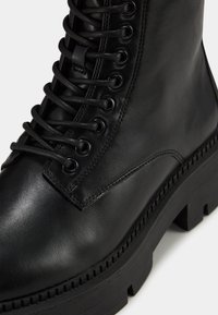 Bottines en cuir noir avec œillets en métal, semelle noire épaisse et lacets bien visibles. Texture lisse avec des détails de couture subtils.