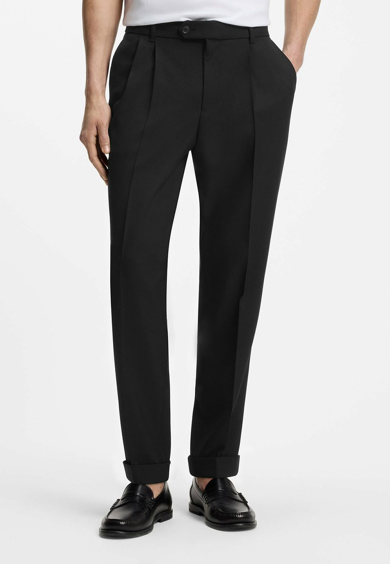 Pantalon noir sur mesure avec poignets, poches latérales et ceinture boutonnée, porté avec des mocassins noirs et une chemise blanche.