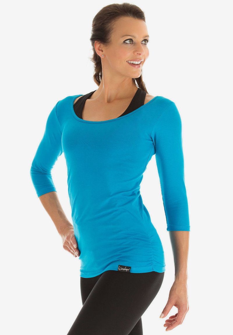 Türkisfarbenes Langarmoberteil mit rundem Ausschnitt und figurbetonendem Design, aus einem dehnbaren Material, das über einem schwarzen Tanktop getragen wird.