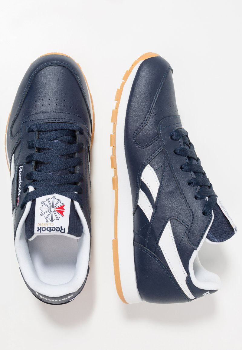 Sneakers in pelle blu navy con dettagli bianchi, superficie texture, punta rotonda, chiusura con lacci e suola in gomma color caramello. Logo Reebok sulla linguetta.