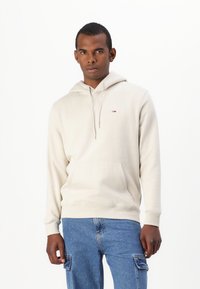 Sudadera beige con un bolsillo frontal tipo pouch, capucha con cordón y un pequeño detalle de logo. Combinada con pantalones cargo de mezclilla azules. Textura suave.