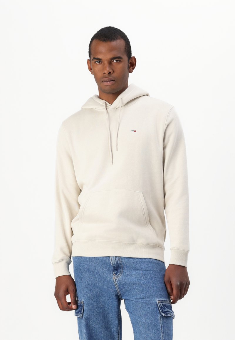 Sudadera beige con un bolsillo frontal tipo pouch, capucha con cordón y un pequeño detalle de logo. Combinada con pantalones cargo de mezclilla azules. Textura suave.