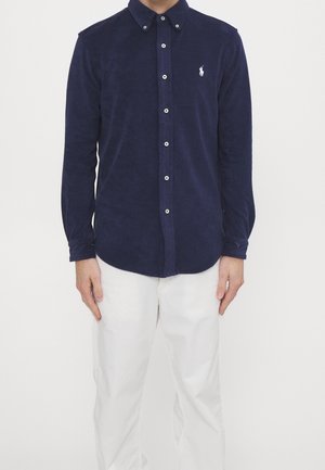 Homme portant une chemise bleu marine à manches longues avec un logo blanc et un pantalon blanc, debout devant un fond clair uni.