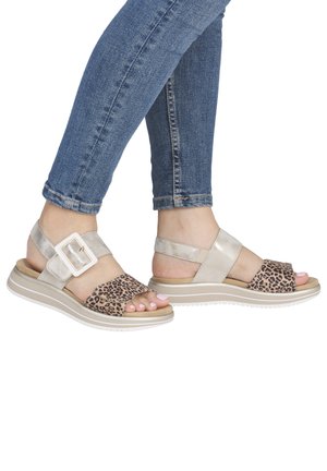 Feet wearing beige and leopard-print sandals with white soles and buckles, paired with blue rolled-up jeans.