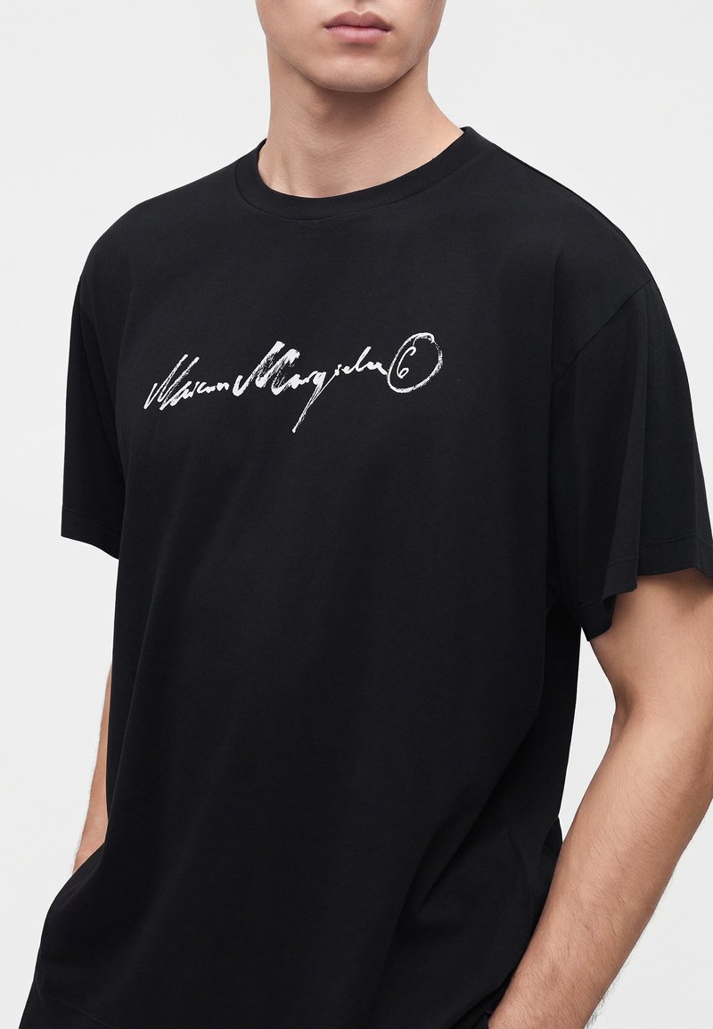 Hombre con camiseta negra con texto blanco en el centro del pecho, manos en los bolsillos, sobre un fondo claro y liso.