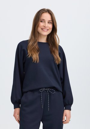 Femme aux longs cheveux bruns souriante, portant un haut à manches longues bleu marine foncé et un pantalon à cordon assorti, debout devant un fond uni.