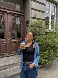 Melns īss topiņš ar džinsa krekla pārnešanu; zilas plaša silueta džinsa bikses. Tur rokā dzērienu ar salmiņu, stāvot pie koka durvīm un zaļumiem.