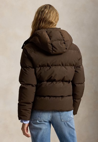 Personne aux longs cheveux portant une doudoune marron foncé avec capuche et un jean bleu clair, vue de dos sur un fond uni.