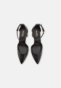 Talons aiguilles noires en cuir avec une fine bride à la cheville et un talon carré de hauteur intermédiaire. Texture lisse et design minimaliste.