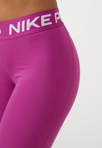 Dopasowane różowe legginsy sportowe z szerokim białym pasem na biodrach z napisem "NIKE PRO". Gładki materiał, tapered design i brak widocznych wzorów.
