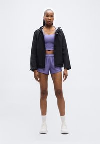 Femme debout en brassière de sport violette et short, avec une veste noire et des baskets blanches, sur un fond blanc uni.