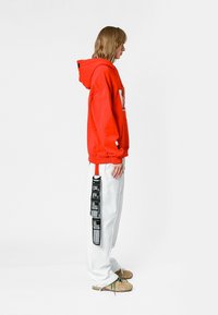 Sudadera roja con capucha y logo en la parte delantera, combinada con pantalones blancos que tienen un detalle de etiqueta negra y cordones verdes en calzado beige.