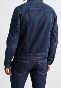 Chaqueta vaquera de mezclilla azul oscuro con corte clásico, puños abotonados y detalle de canesú en la espalda, combinada con jeans oscuros ajustados a juego.