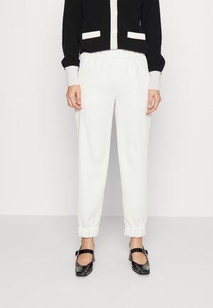 Personne portant un pantalon blanc à taille élastique et des chaussures Mary Jane noires avec un haut à manches longues noir et blanc, debout devant un fond uni.