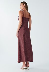 Žena s dlhými vlasmi, oblečená v hnedej šaty s dĺžkou po členky, posiatymi malými bielymi bodkami a bielymi sandálmi na podpätku, stojaca bokom proti jednoduchému pozadiu.