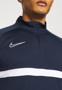 Granatowy sportowy top na zamek błyskawiczny z białym paskiem i małym logo Nike. Materiał wydaje się gładki o lekkiej teksturze.
