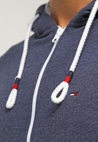 Navyblå zip-up hoodie med vita repdragkedjor och röda och svarta accenter, som bärs på en persons övre bröst och nacke.