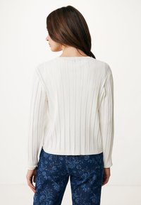 Biały sweter z dzianiny, z pionowym prążkowaniem, z długimi rękawami i szerokim dekoltem. W zestawie z niebieskimi spodniami w wzory.