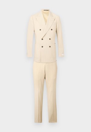 Blazer cruzado color crema con botones marrones combinado con pantalones a medida a juego sobre un fondo claro y sencillo.