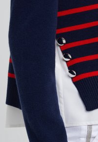 Navy sweater with red horizontal stripes, featuring three silver buttons on the side and ribbed texture on the sleeve.