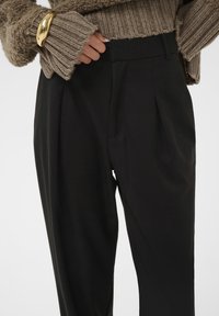 Crne krojene hlače s pregibima i bočnim džepovima, uparene s teksturiranim bež pletenim puloverom s rebrastim rukavima i zlatnom narukvicom.