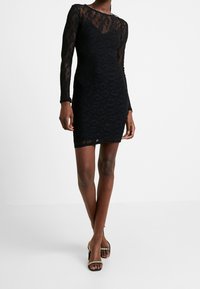 Femme portant une robe noire à manches longues en dentelle avec un motif animalier et des sandales à talons noires avec des brides en strass.