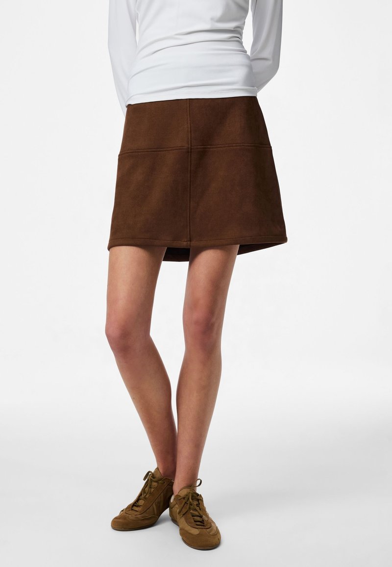 Femme portant une mini-jupe marron évasée avec des coutures visibles, des baskets vert olive et un haut blanc à manches longues, debout jambes croisées.