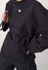 Persona che indossa un maglione nero a maniche lunghe con una piccola toppa del logo, con i lacci in vita annodati, che tiene una tracolla nera testurizzata sulla spalla.
