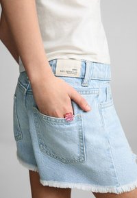 Denim shorts in light blue, with a frayed hem. A pink flower ring is worn on the hand, tucked into the side pocket. Tag on shorts reads "MNG KIDS DENIM LAB."