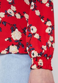 Blouse florale rouge avec des roses blanches et des feuilles bleues. Dotée de poignets élastiques et d'une texture lisse. Portée sur un jean bleu clair.
