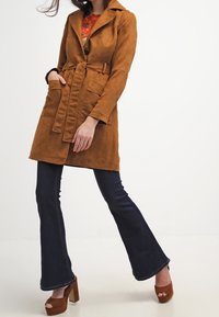 Cappotto trench in suede marrone con vita regolabile, colletto a punte e due tasche frontali, abbinato a jeans svasati scuri e tacchi a pianta larga.