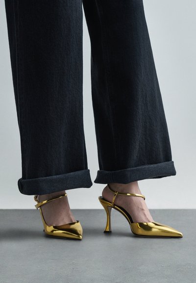 Talons aiguilles dorés à bout pointu et à lanière, au design épuré avec un fin talon. Portés avec un pantalon en denim foncé retroussé.
