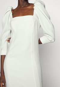 Robe blanche ajustée avec un décolleté carré, des manches bouffantes et une texture lisse ; présente des coutures structurées sur les côtés.