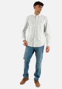 Camicia a maniche lunghe, a strisce blu chiaro, con colletto button-down, indossata con jeans blu e scarpe marroni. Il tessuto appare semplice e informale.