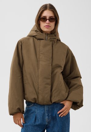 Kvinde iført brun oversized dunjakke med hætte, blå jeans og små rektangulære solbriller, står mod en ensartet lys baggrund.